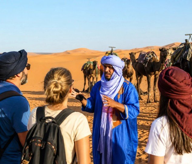 First One Travel guide with travelers among camels in the Sahara